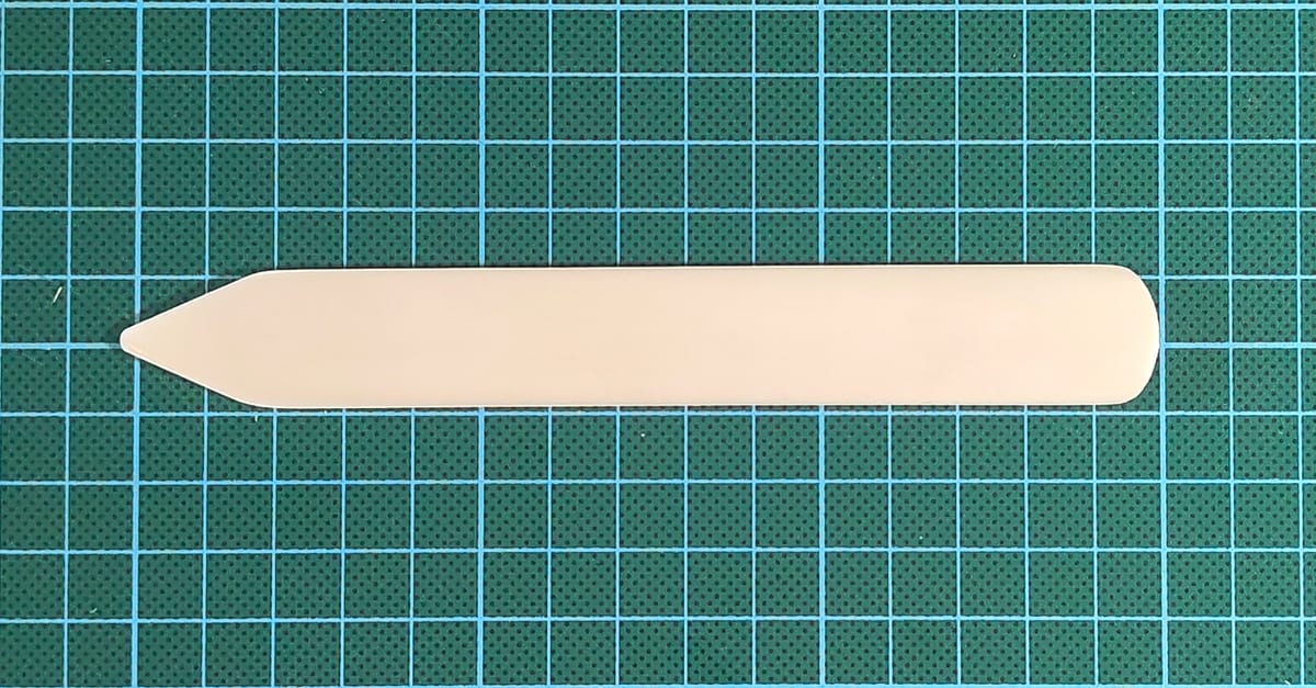 A pointed plastic bone folder lying on a green cutting mat.
