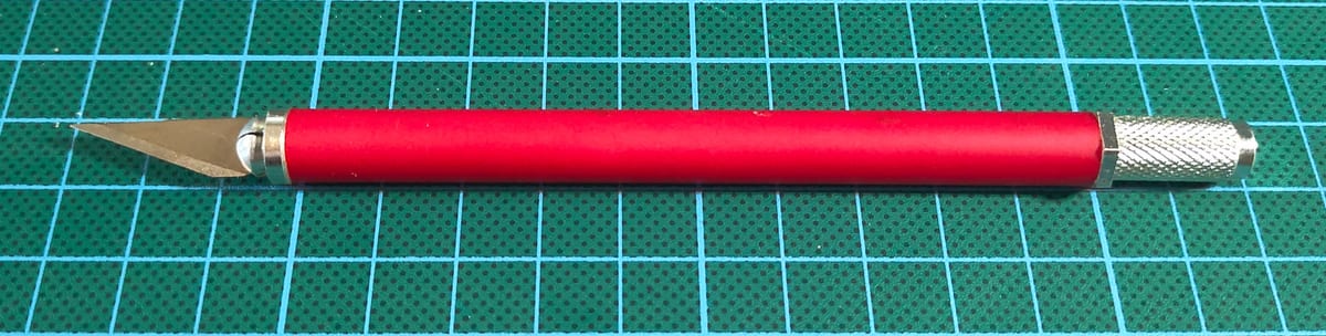 A red-handled craft knife with a triangular blade on a green cutting mat.