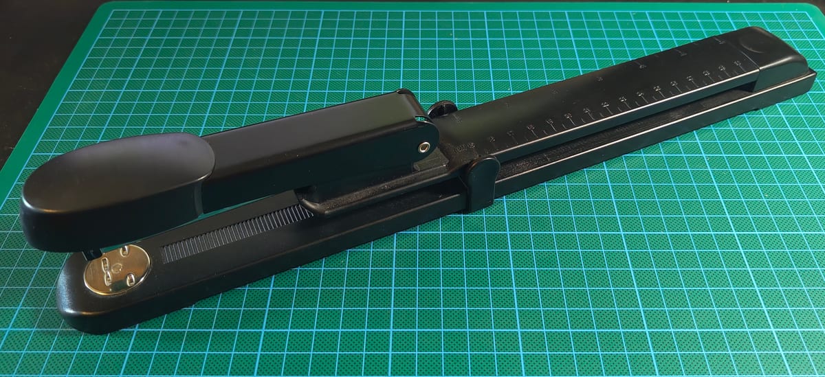 A black long-arm stapler on a green cutting mat, with the scale markings along its extended arm.